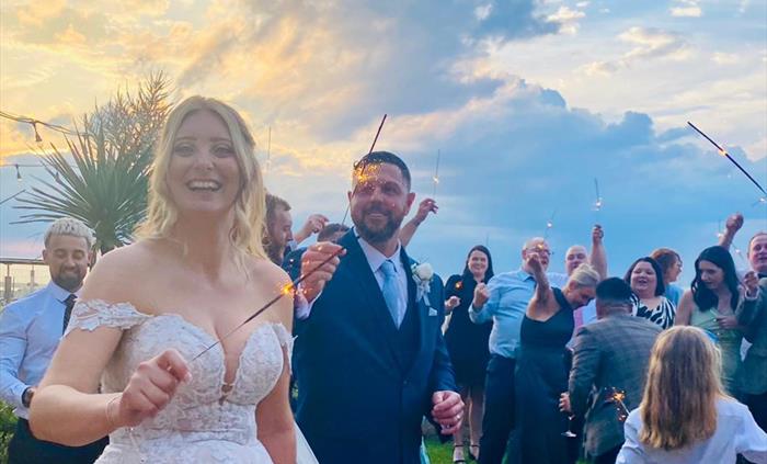 An image of a newly wedded bride and groom holding sparklers with their guests with the beach in the background, and the clouds golden from the sunset